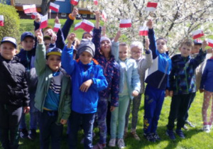 Dzieci pozują do zdjęcia z małymi flagami na tle kwitnącego drzewa.